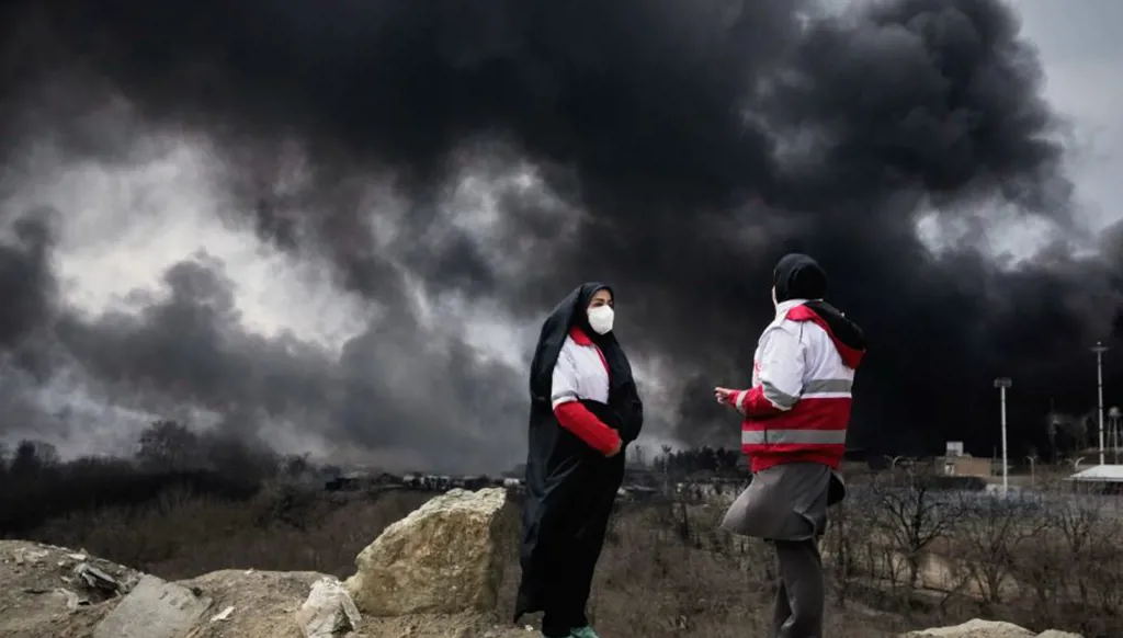 امرأتان من جمعية الهلال الأحمر الإيراني تقفان بينما يتصاعد عمود كثيف من الدخان في السماء في طهران، (أ ب).
