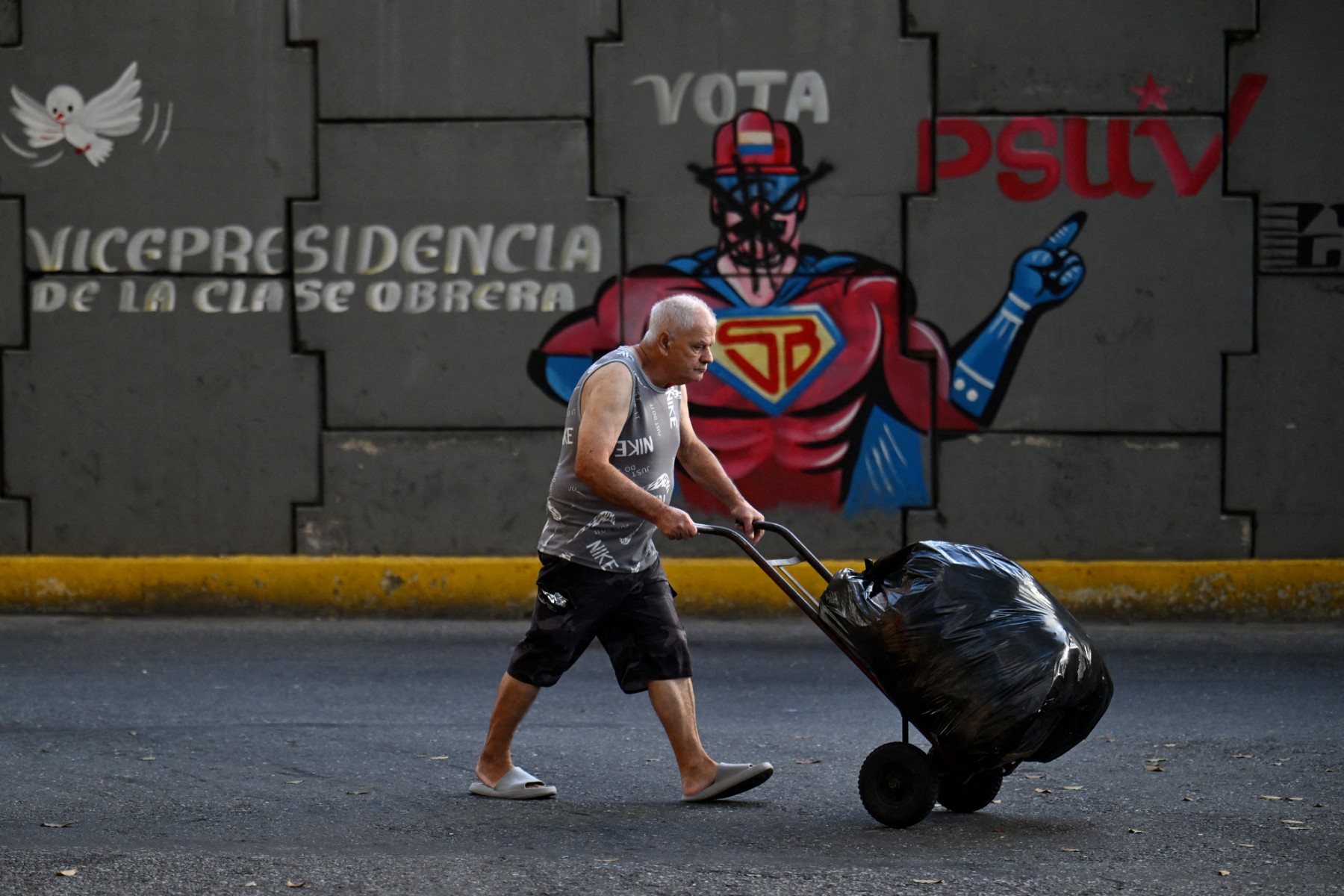رجل يحمل عربة يمر أمام جدارية تصور الرئيس الفنزويلي المخلوع نيكولاس مادورو بشخصية تدعى ”سوبر-بيغوتي“ (أ ف ب).