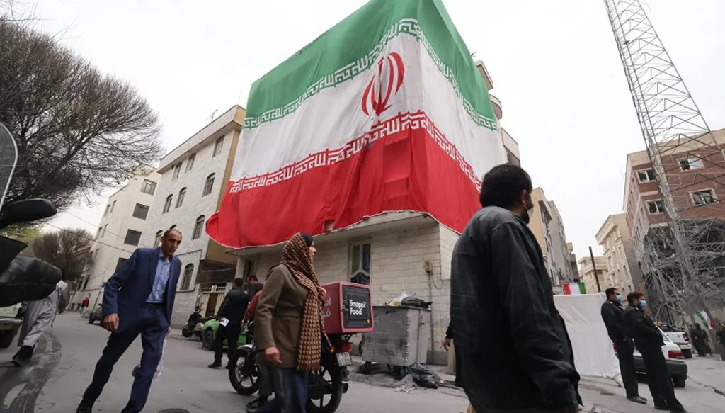 علم إيراني ضخم على مبنى بجوار مباني متضررة في أعقاب غارة عسكرية سابقة في طهران. (أ ف ب).