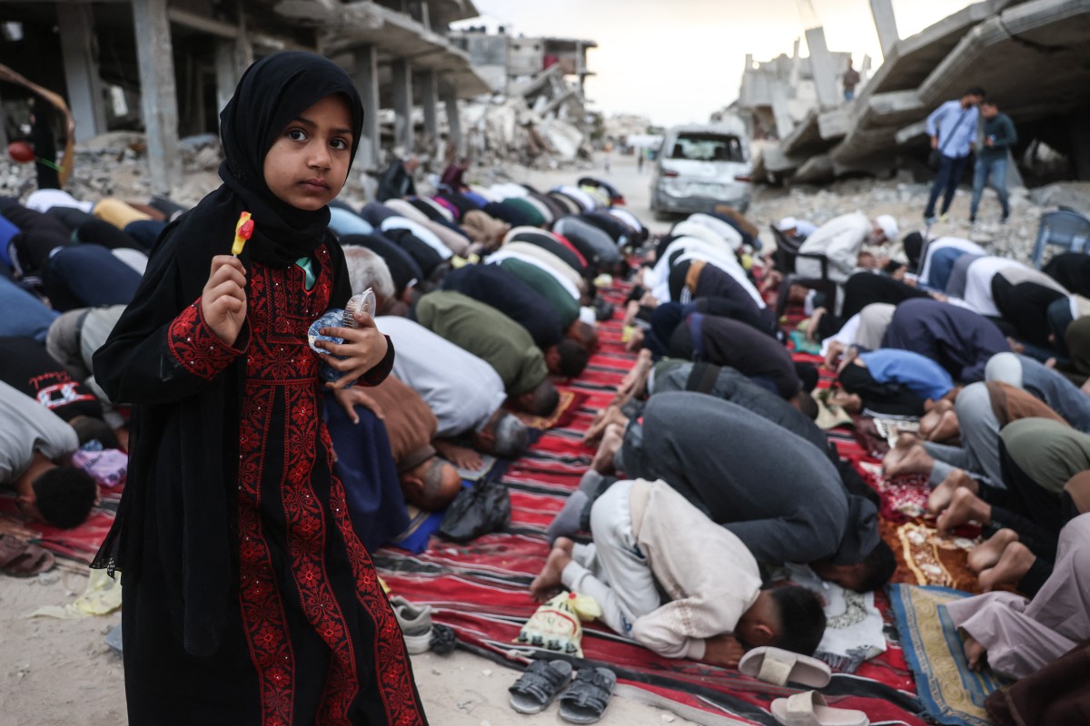 فتاة تحمل قطعة حلوى تقف بين الفلسطينيين الذين يؤدون صلاة الصباح بمناسبة بداية عيد الأضحى، في شارع متضرر بشدة في خان يونس