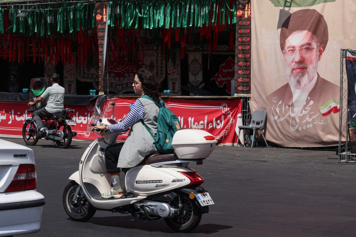 سيدة تقود دراجة نارية أمام لافتة تحمل صورة المرشد الإيراني مجتبى خامنئي في ساحة ولي عصر في طهران. (أ ف ب)