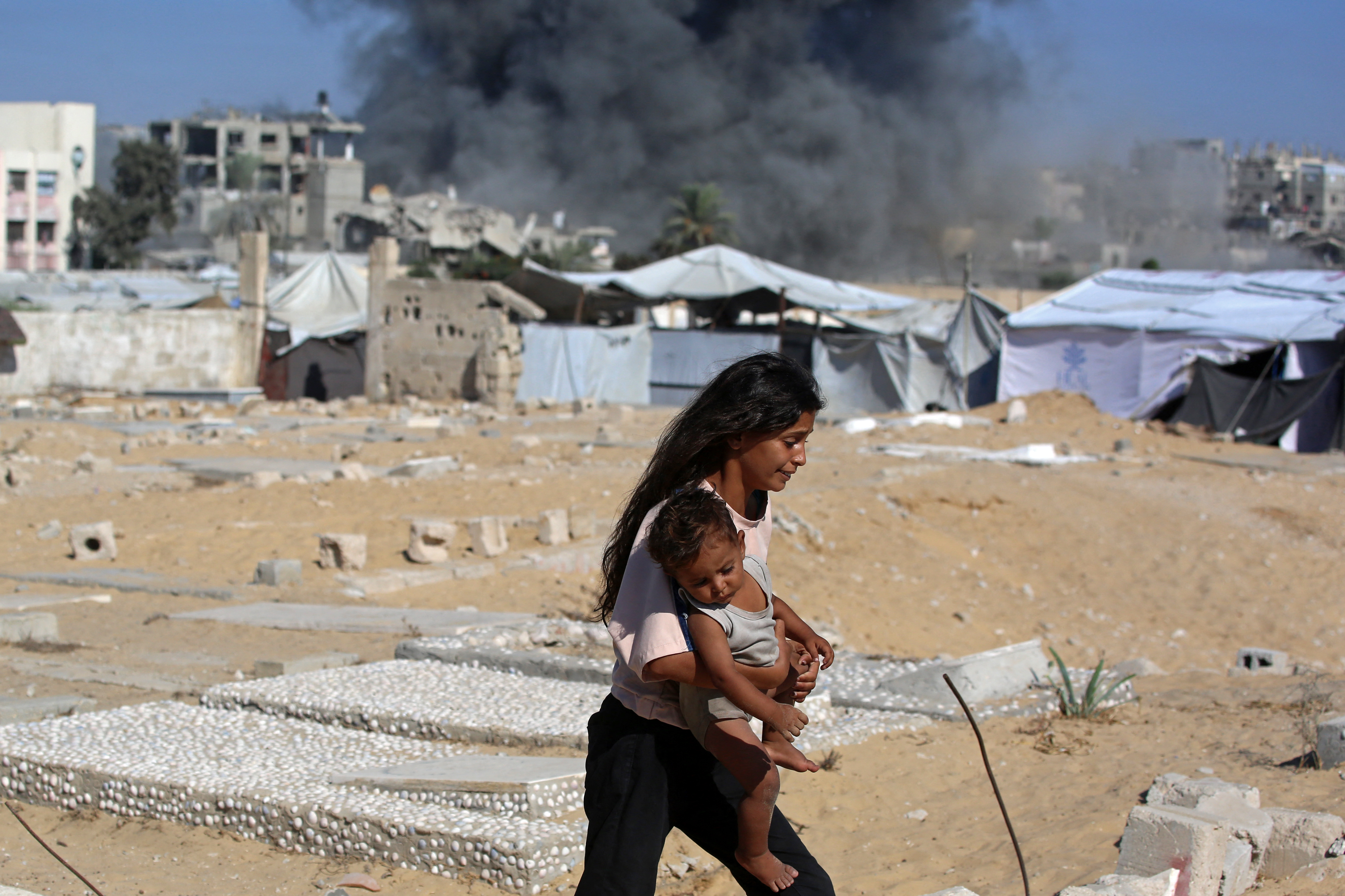 فتاة فلسطينية تحمل طفلاً وفي الصورة يظهر منزل استُهدف بغارة إسرائيلية في خان يونس في 9 سبتمبر 2026 (أ ف ب)