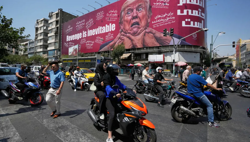 سائقو دراجات نارية يمرون أمام لوحة إعلانية تُظهر يدين تطوقان عنق الرئيس الأميركي دونالد ترامب، وتحمل عبارة "الانتقام حتمي"، في طهران في 24 آب/أغسطس 2026. (أ ب)