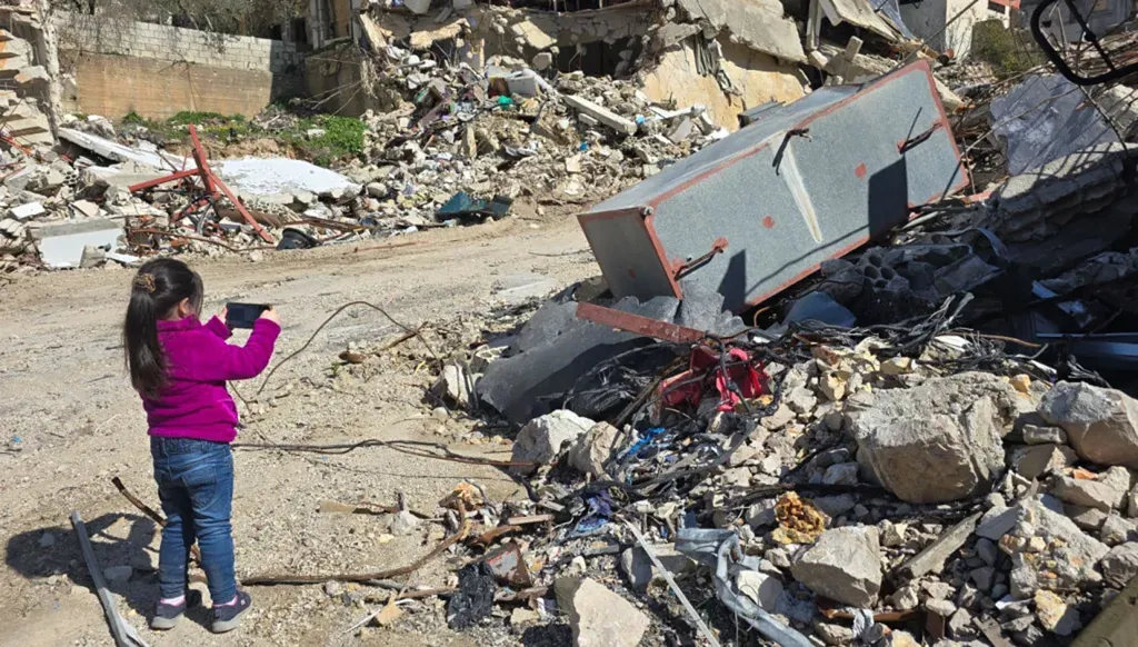 طفلة تلتقط صورة لمنزل والدها المدمّر في بلدة عديسة الحدودية جنوبي لبنان (تصوير أحمد منتش).