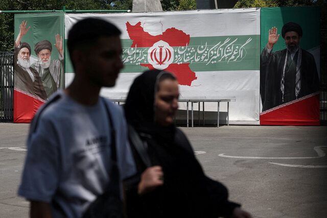 شخصان يمران أمام لافتة عليها صور الزعيم الراحل للثورة الإسلامية آية الله روح الله الخميني، والزعيم الأعلى الراحل لإيران آية الله علي خامنئي، والزعيم الأعلى الحالي لإيران مجتبى خامنئي، في أحد شوارع طهران. (رويترز)