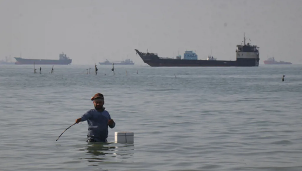A man stands in the water, appearing to fish, as bulk carriers, cargo ships, and service vessels line the horizon in the Strait of Hormuz off Bandar Abbas, Iran, Monday, April 27, 2026.(Razieh Poudat/ISNA via AP)