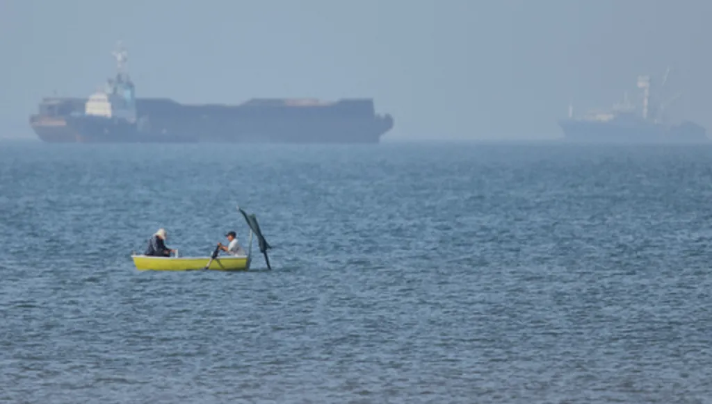 Two men sit in a small boat on the water as cargo ships are anchored in the background in the Strait of Hormuz off Bandar Abbas, Iran, Monday, May 4, 2026. (Amirhosein Khorgooi/ISNA via AP)