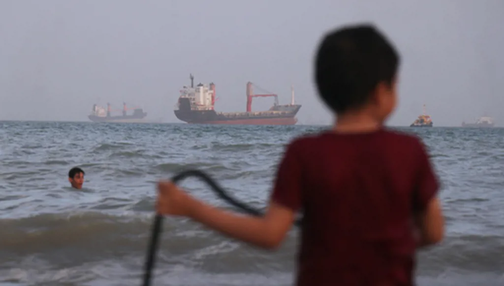 Children play in the water along the shore as a mix of bulk carriers, cargo ships, and service vessels sit offshore in the Strait of Hormuz off Bandar Abbas, Iran, Sunday, April 26, 2026.(Razieh Poudat/ISNA via AP)