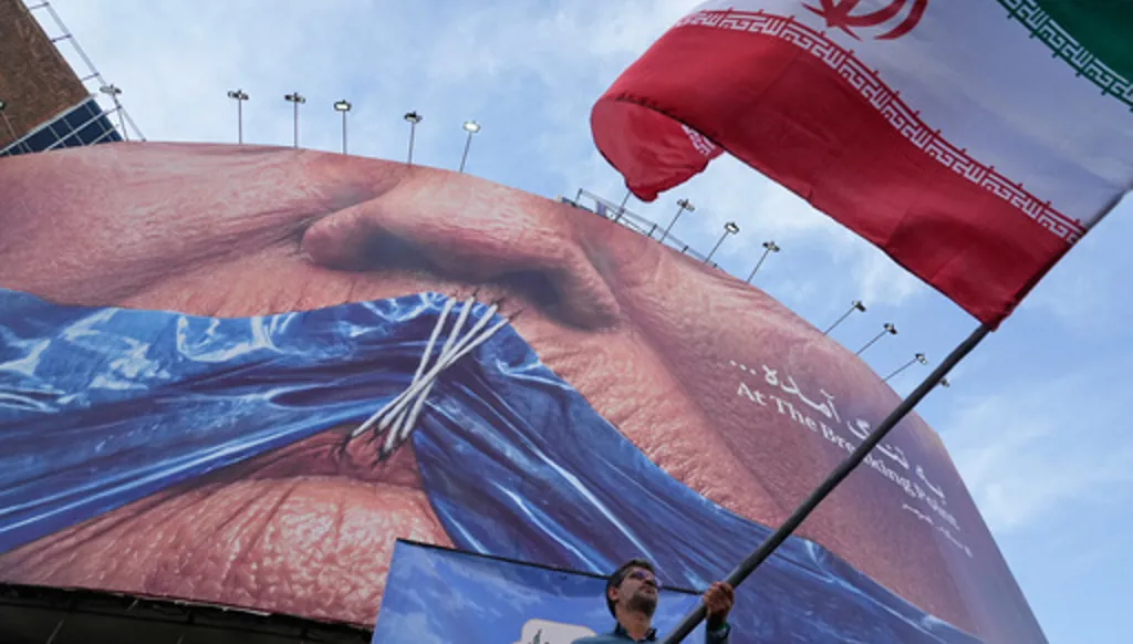A man waves an Iranian flag for a pro-government campaign under a billboard with graphic showing Strait of Hormuz and sewn lips of U.S. President Donald Trump in a square in downtown Tehran, Iran, Wednesday, May 6, 2026. (AP Photo/Vahid Salemi)