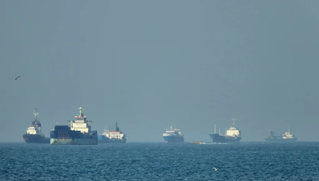 Cargo ships, including bulk carriers and general cargo vessels, sit at anchor offshore as a small motorboat passes in the foreground, in the Strait of Hormuz off Bandar Abbas, Iran, Monday, May 4 , 2026.(Amirhosein Khorgooi/ISNA via AP)