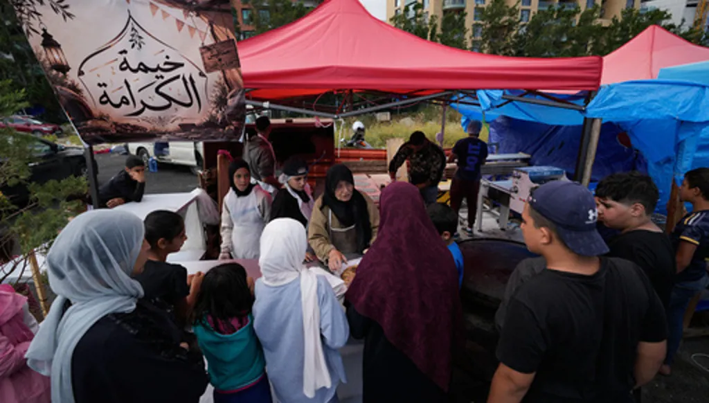 Soubhiye Zeiter, 63, centre, a displaced woman who fled her home with her family in Beirut's southern suburbs, speaks with displaced people who lined up to receive mana'eesh, a popular Lebanese flatbread breakfast topped with thyme, at a small bakery in a tented settlement along Beirut's waterfront, Lebanon, Thursday, May 21, 2026. The Arabic words on the top left read:"The tent of dignity." (AP Photo/Hussein Malla)