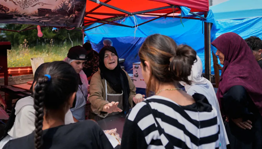 Soubhiye Zeiter, 63, centre, a displaced woman who fled her home with her family in Beirut's southern suburbs due to the war between Israel and Hezbollah, speaks with displaced people who lined up to receive mana'eesh, a popular Lebanese flatbread breakfast topped with thyme, at a small bakery in a tented settlement along Beirut's waterfront, Lebanon, Thursday, May 21, 2026. (AP Photo/Hussein Malla)