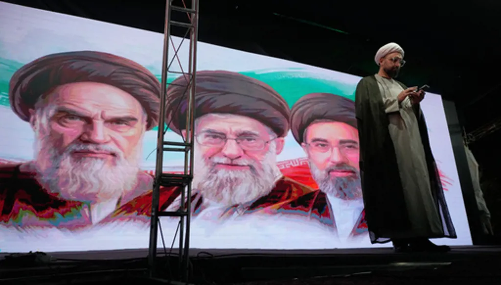 A cleric checks his cell phone on stage in front of a screen displaying portraits of the late Iranian revolutionary founder Ayatollah Khomeini, left, late Supreme Leader Ayatollah Ali Khamenei, center, and Supreme Leader Ayatollah Mojtaba Khamenei, during a pro-government gathering in Tehran, Iran, Sunday, June 7, 2026. (AP Photo/Vahid Salemi)