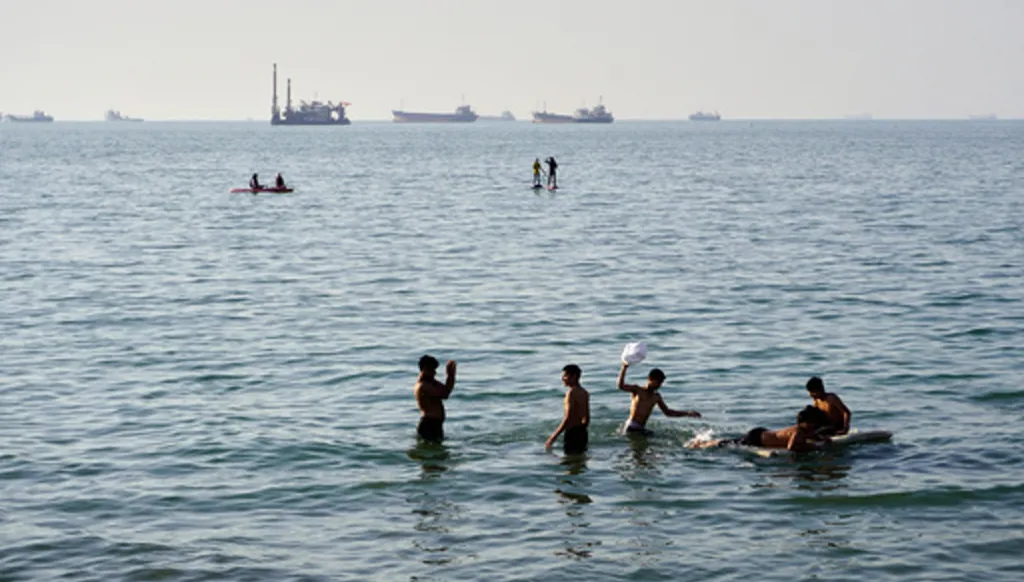 Residents swim and play in the waters of the Strait of Hormuz while cargo ships and commercial vessels lie anchored in the distance off Bandar Abbas, Iran, Wednesday, June 10, 2026.(Razieh Poudat/ISNA via AP)