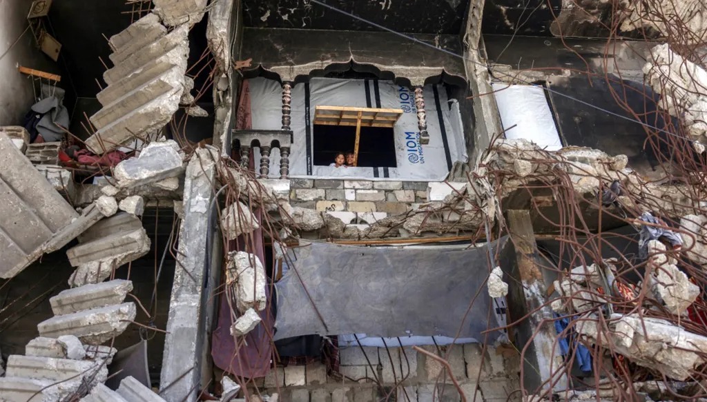 طفلان يطلّان من نافذة مأوى موقت وسط أنقاض مبنى متضرر بشدة في خان يونس جنوب قطاع غزة، 25 آب/أغسطس 2026. (أ ف ب)
