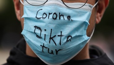 رجل يرتدي قناع وجه مكتوب عليه "دكتاتورية كورونا" في فرانكفورت أم ماين، غرب ألمانيا، احتجاجاً على القيود المفروضة لمكافحة الوباء (2 أيار 2020 - أ ف ب).