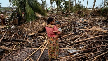 امرأة من الروهينغا تحمل طفلها بجوار منزلها المدمّر في مخيم بسارة للاجئين في سيتوي في بورما بعدما ضرب إعصار موكا اليابسة (16 أيار 2023، أ ف ب). 