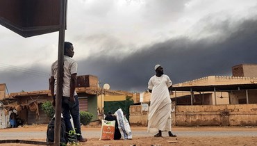 دخان يتصاعد فوق المباني بينما ينتظر سودانيون على جانب طريق مع أغراضهم، في الخرطوم (10 حزيران 2023 - أ ف ب).