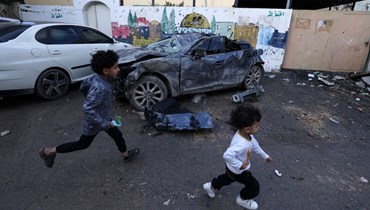 أطفال فلسطينيّون يمرّون بجوار سيّارة متضررة بعد غارة شنّها الجيش الإسرائيلية في الصباح الباكر، في مخيم جنين بالضفة الغربية المحتلّة (أ ف ب). 
